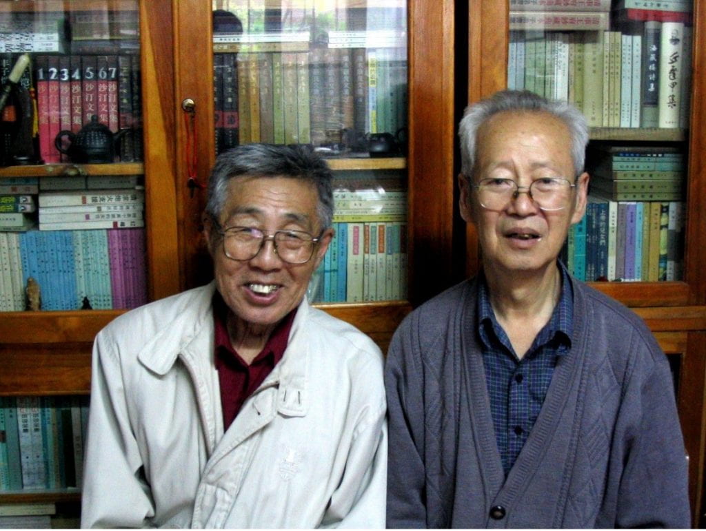 two elderly men sitting beside one another and smiling infront of a bookshelf