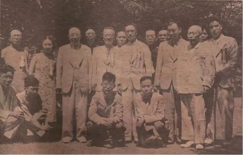 A group of people (17 visible) stand together in front of a backdrop of trees.