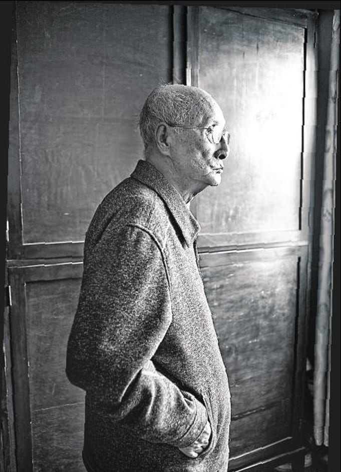 An elderly man stands with his hands in his pockets, looking off-image