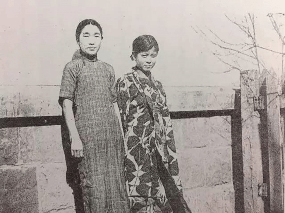 black and white photograph of two young women smiling and posing outside along a fence and trees