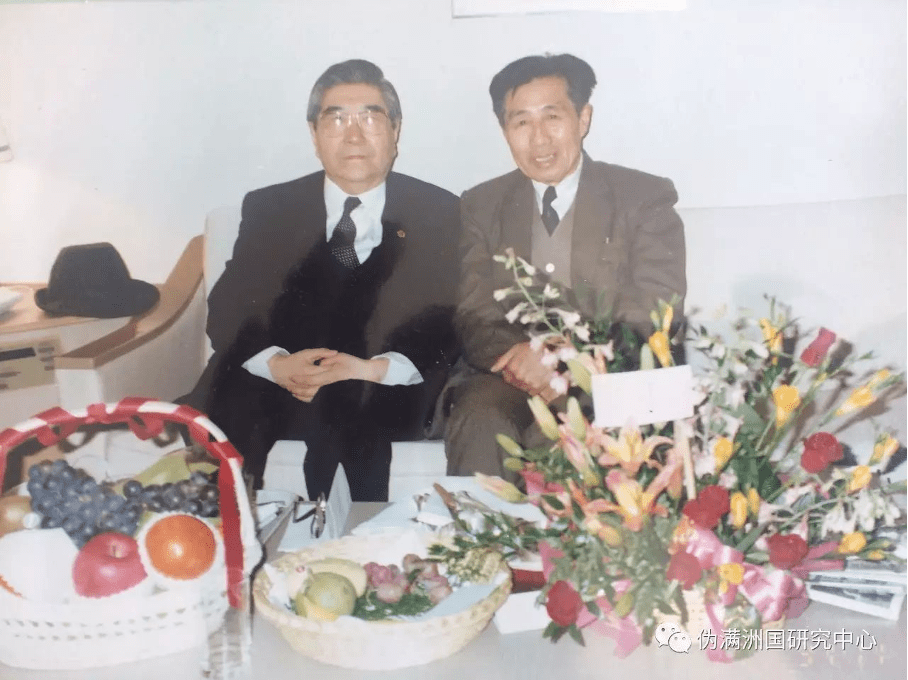 photograph of two elderly men sitting beside one another behind a table of food and flowers