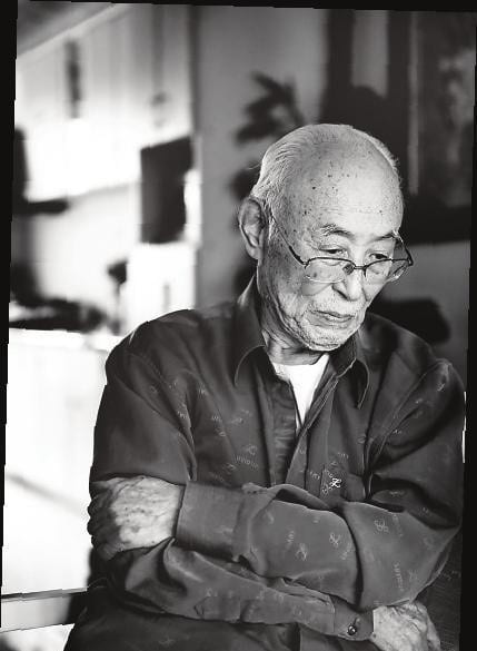 black and white photograph of an elderly man with glasses staring at the ground with his arms crossed