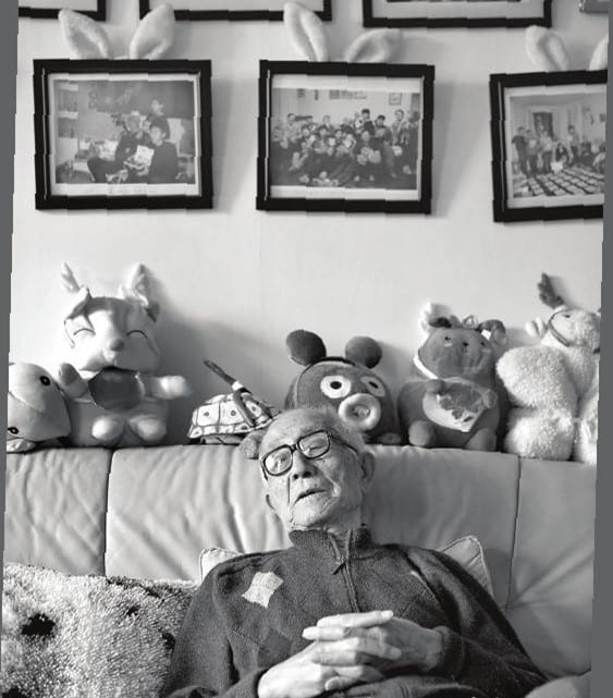 black and white photograph of an elderly man resting on a couch with stuffed toys and framed pictures behind him